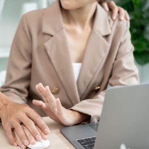 A man and woman are seated at a desk, clasping hands in a gesture of shock.
